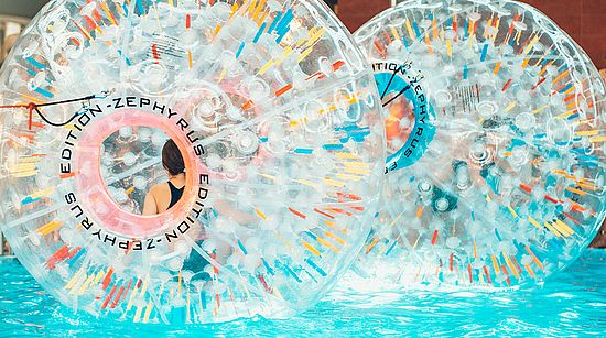 Zwei transparente aufblasbare Wasserlaufbälle mit farbigen Griffen in einem Schwimmbecken, eine Person im schwarzen Badeanzug ist teilweise sichtbar