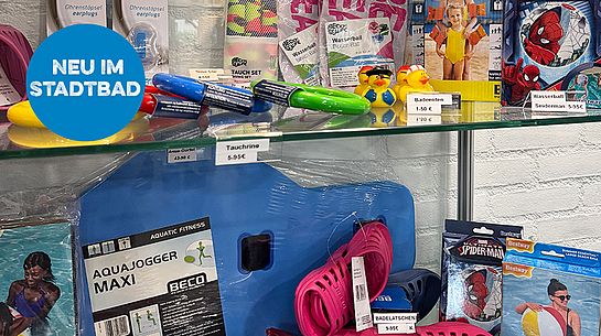 Vitrine im Stadtbad Schwerte mit verschiedenen Schwimmutensilien wie Badeenten, Tauchringen, Schwimmbrettern, Badelatschen und Wasserbällen. Ein blauer Störer mit der Aufschrift „Neu im Stadtbad“ weist auf das neue Angebot hin.