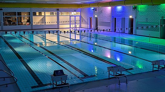 Ein bunt beleuchtetes Schwimmbecken im Stadtbad Schwerte. Eine Girlande mit kleinen Fähnchen ist über das leere Schwimmbecken gespannt.  