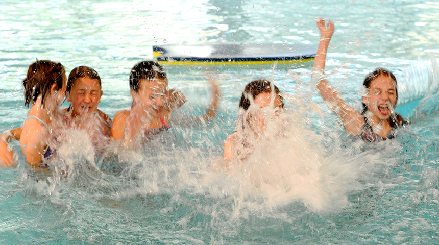 Eine Gruppe Kinder im Wasser, im Vordergrund spritzt viel Wasser nach oben, der Hintergrund ist unscharf. 