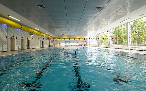 Innenansicht des Stadtbad Schwerte. Zu sehen ist das große 25m-Schwimmbecken mit mehreren Schwimmern im Wasser und großen Fenstern rechts